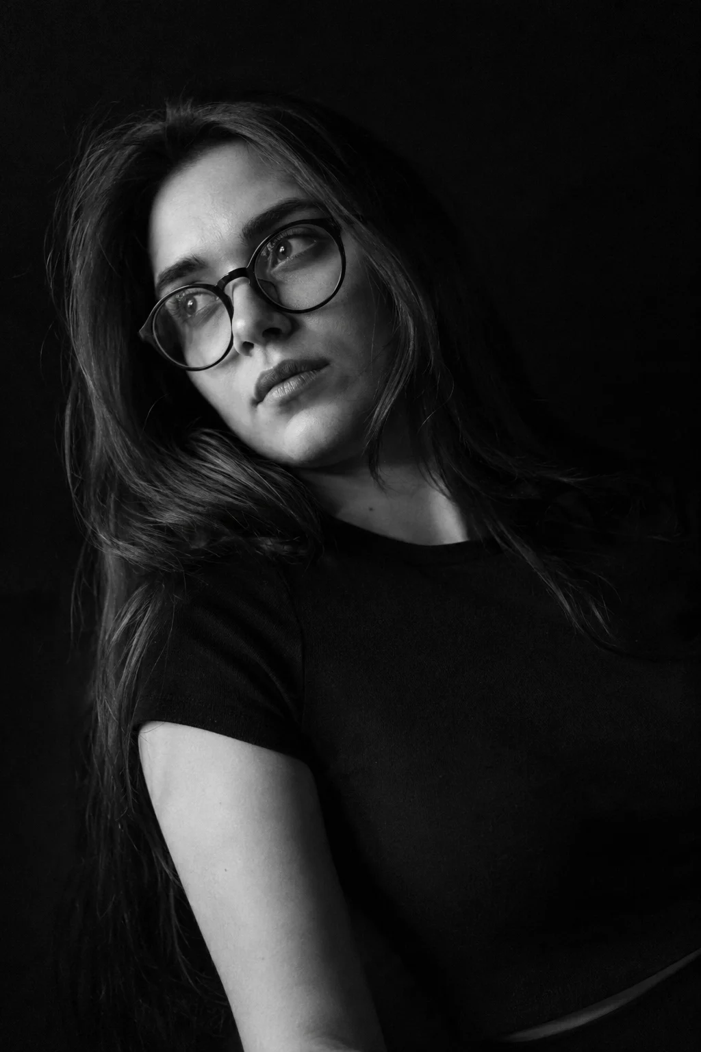 Black and white smartphone portrait of a young woman with round glasses leaning diagonally in a dark indoor setting.