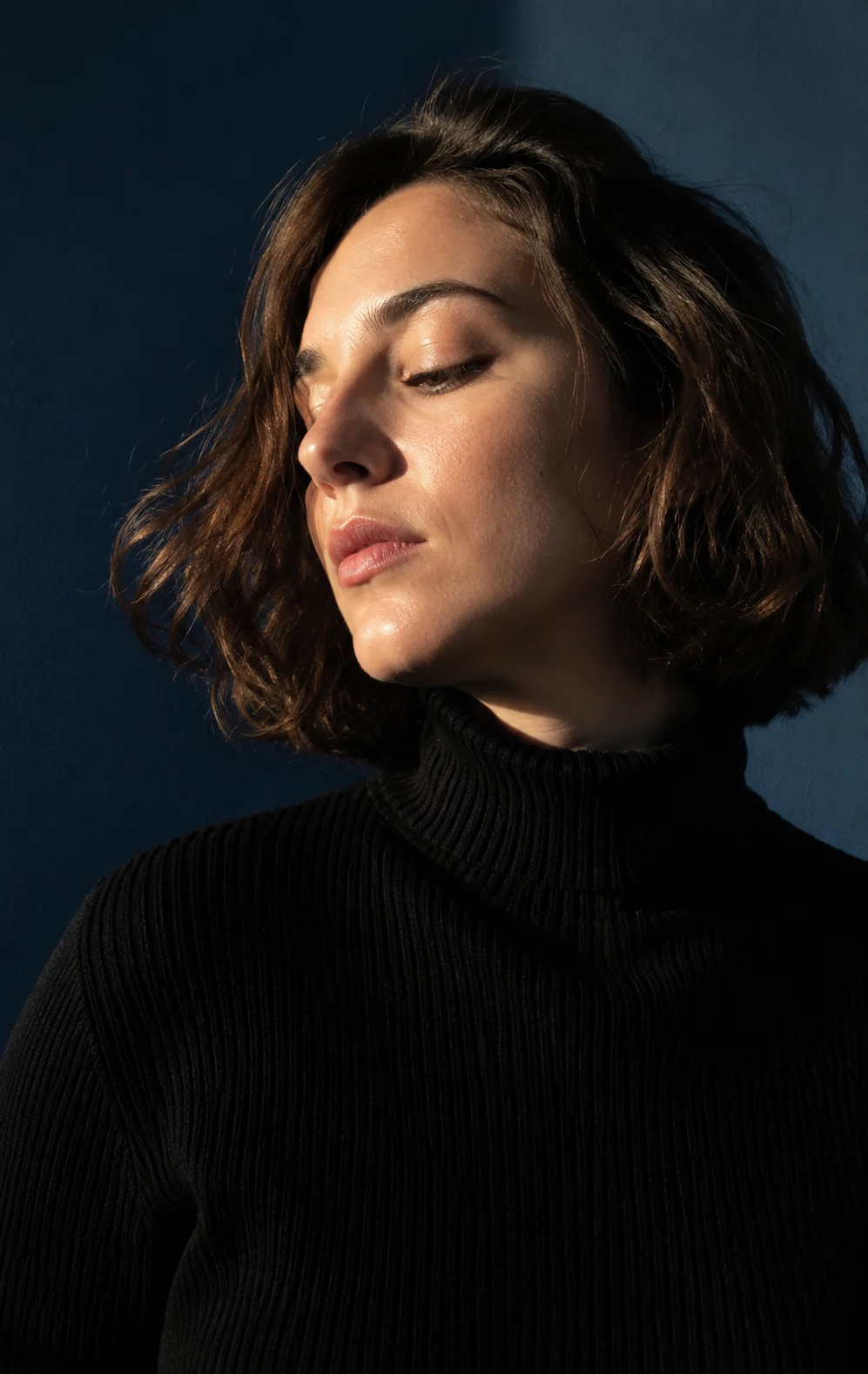 Vertical smartphone portrait of a young brunette woman with closed eyes, dark turtleneck, warm sunlight on face, deep teal wall background.