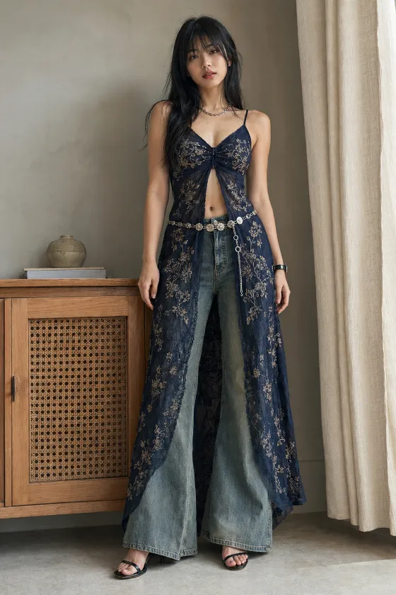 Full-body fashion styling with navy embroidered lace layered over wide-leg faded denim, silver floral belt, and open-toe heels in a softly lit neutral interior.