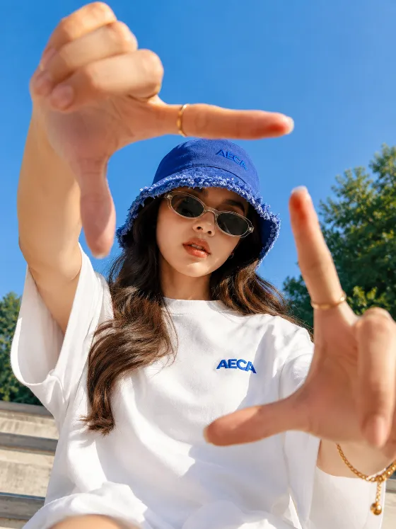 Outdoor editorial portrait of a fashion subject wearing a blue bucket hat and oversized white T-shirt while framing the camera with extended hands.