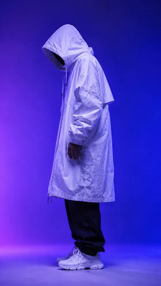 Full-body side profile of a hooded model in an oversized white techwear coat against a purple-blue studio gradient.