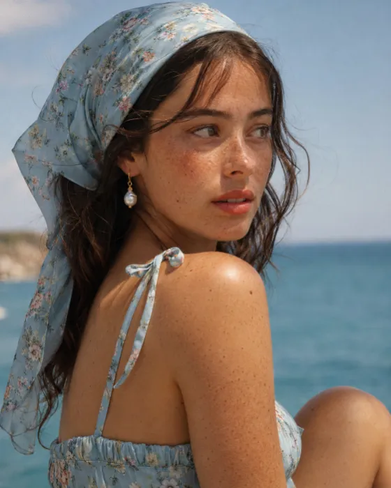 Close coastal fashion portrait of a woman in a pale blue floral headscarf and matching strappy top, with a pearl-like drop earring and sunlit freckles against a blurred blue sea background.