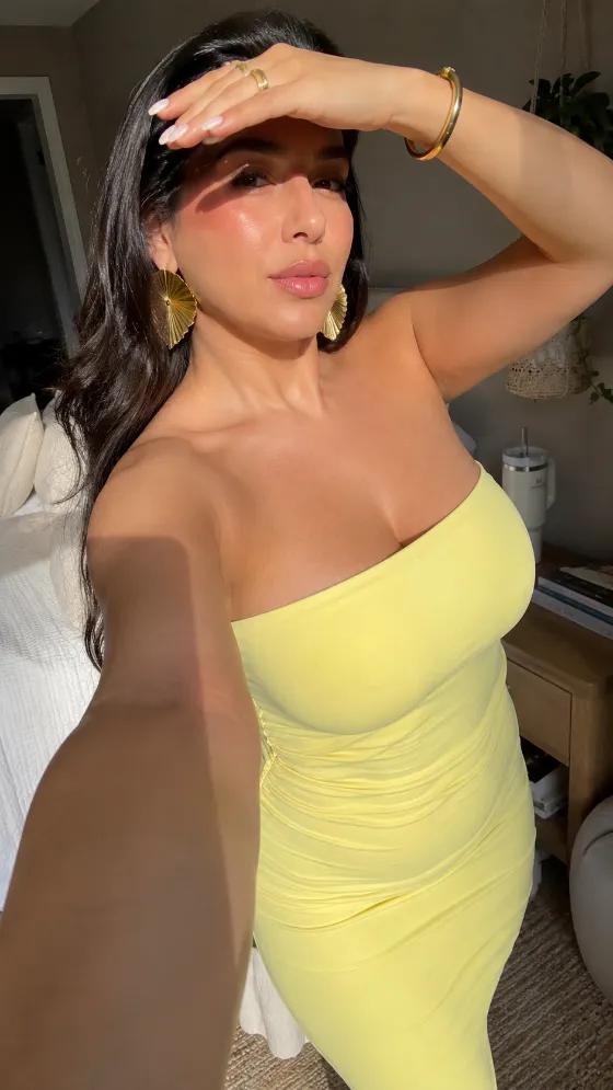 Sunlit woman wearing a pale yellow strapless bodycon dress with gold fan earrings in a bedroom