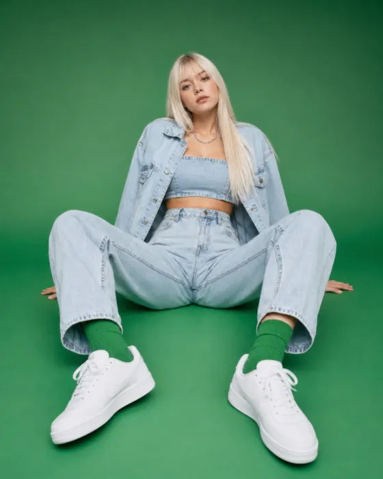 Studio fashion portrait of a platinum-haired subject seated on a green seamless backdrop wearing a denim jacket, cropped denim top, and loose jeans.