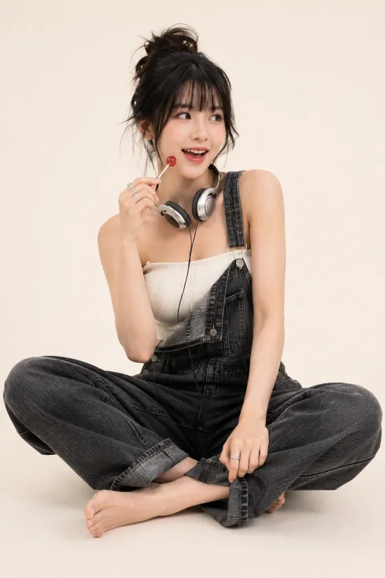 Vertical casual denim editorial with overalls, headphones, a lollipop, bare feet, and a playful seated pose.