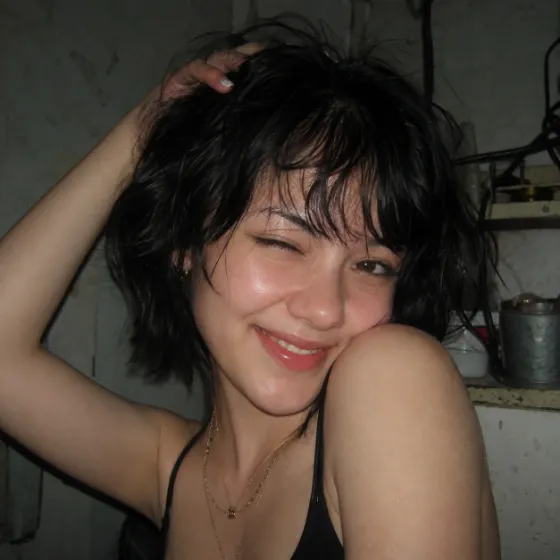 Close-up of a winking woman with a tousled black bob, black top, and layered gold necklaces under harsh direct flash in a cluttered dim room.