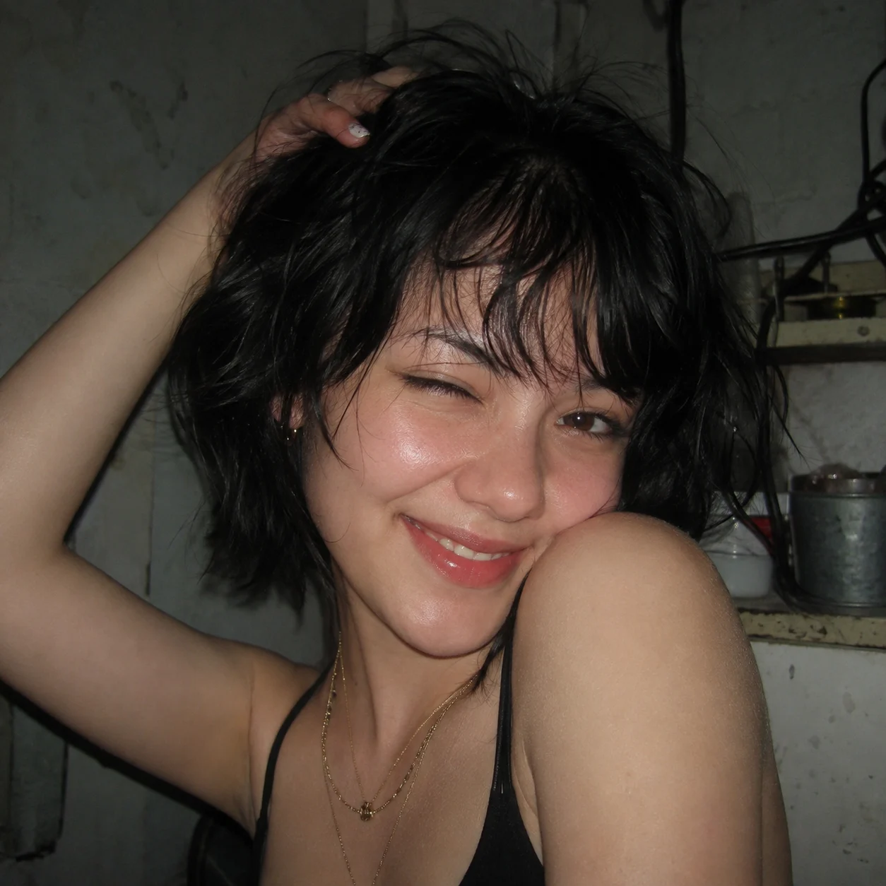 Close-up of a winking woman with a tousled black bob, black top, and layered gold necklaces under harsh direct flash in a cluttered dim room.