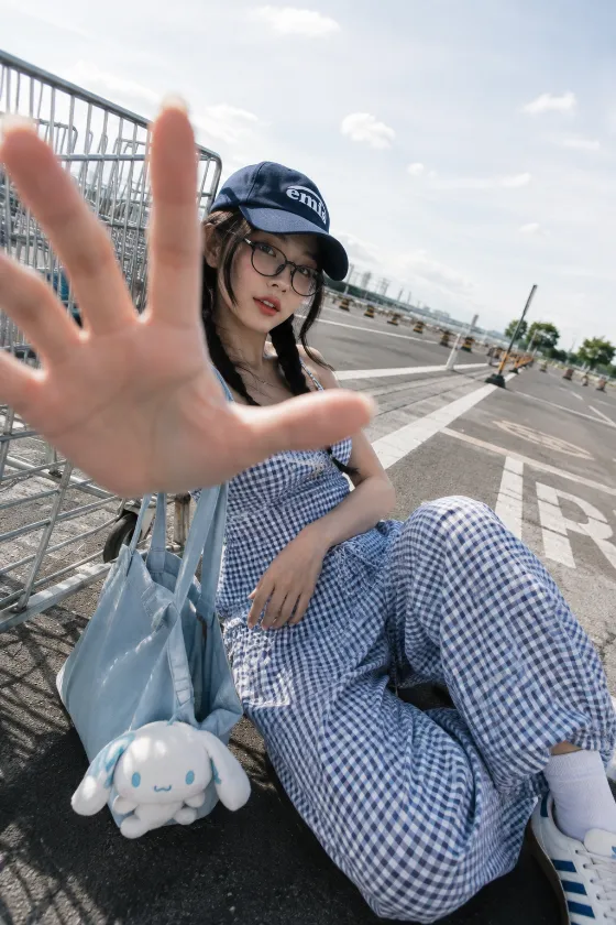 Playful gingham street-fashion portrait with cap, round glasses, plush tote, and foreground hand