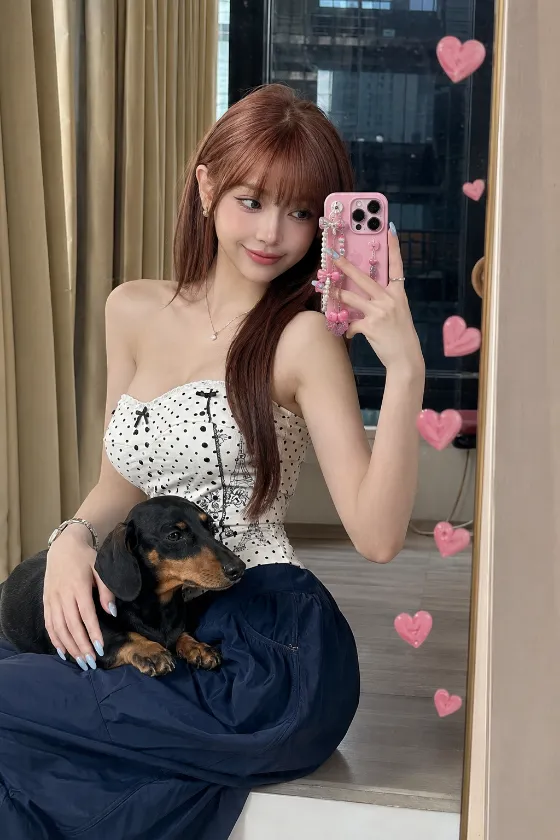 Playful mirror-selfie fashion portrait with a polka-dot camisole, pink phone accessories, beige curtains and a dachshund companion.