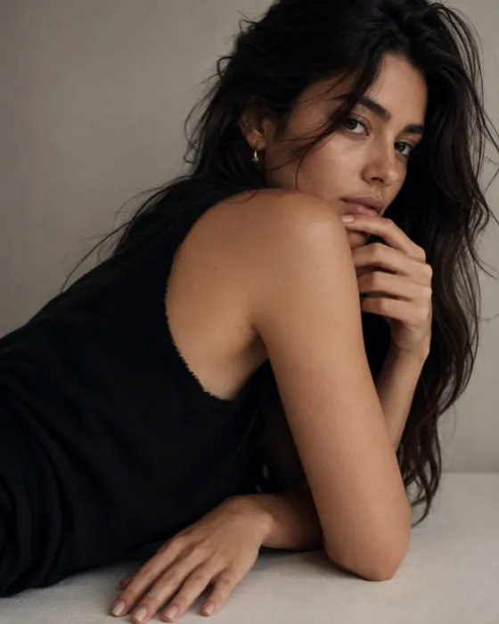 Minimal editorial portrait with a reclined over-shoulder pose, matte black top, pale surface, and warm-gray studio background.