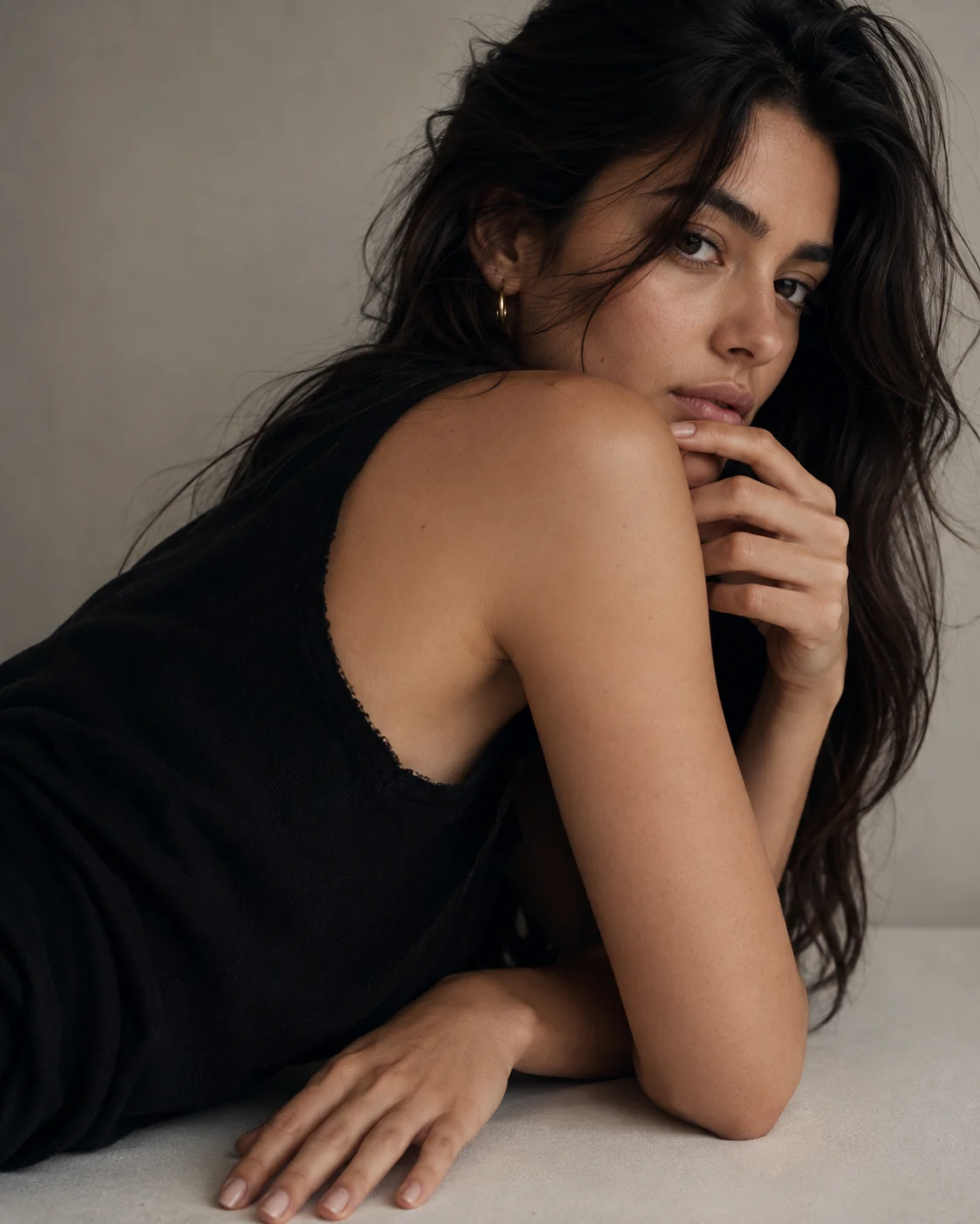 Minimal editorial portrait with a reclined over-shoulder pose, matte black top, pale surface, and warm-gray studio background.