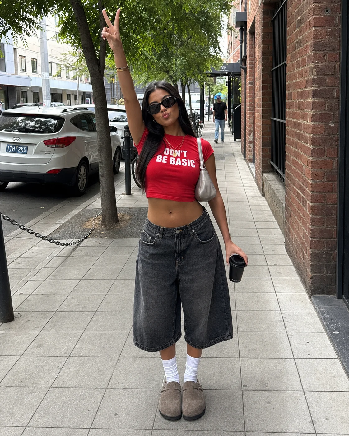 Street-style outfit with a red cropped graphic tee, wide charcoal denim culottes, sunglasses, socks, and chunky shoes on a city sidewalk.