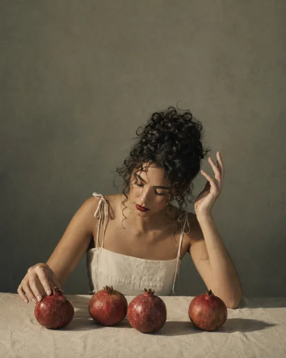 Fashion subject with dark curly hair leaning over four pomegranates on a cream table under hard directional studio light.