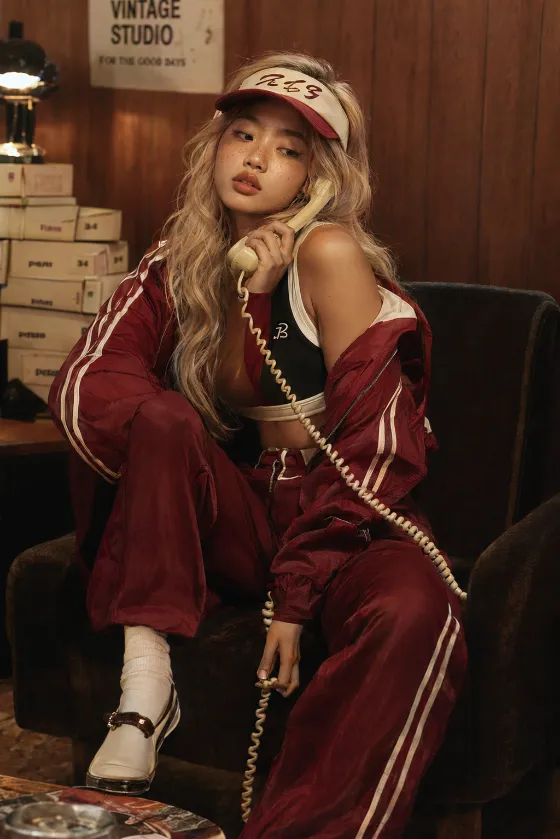 Seated streetwear portrait with burgundy track layers, a visor cap and cream telephone against a warm wood-paneled interior.