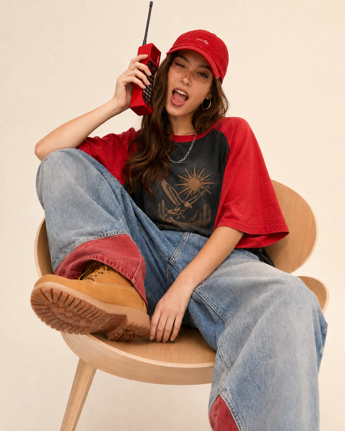 Seated street-fashion portrait of one woman in oversized graphic streetwear, baggy denim, tan boots, and a red cap while holding a red brick-style phone.