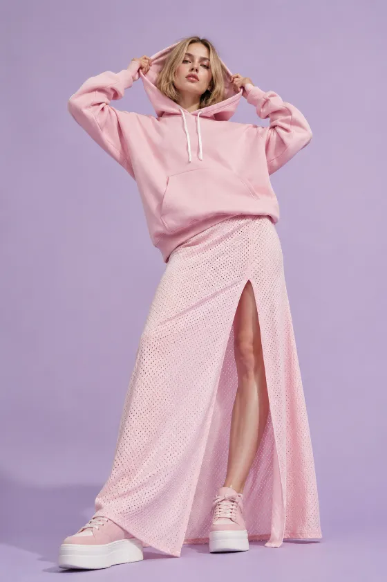 Young Scandinavian fashion subject with porcelain skin and a chin-length bob hairstyle posing in a pink hoodie and matching slit skirt against a lilac studio background, with both hands holding the hood edges in a low-angle editorial pose.