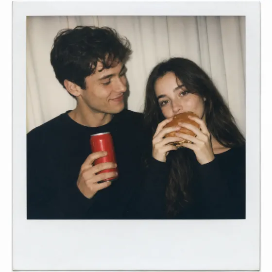 Young couple in black tops, one holding a red soda can, the other a burger near her mouth, pale curtain background, instant-photo style.