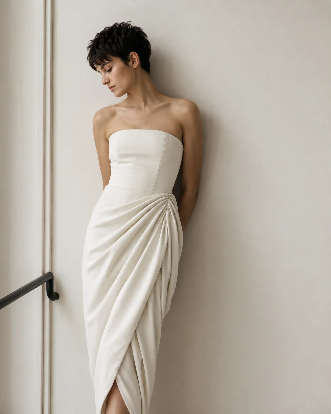 Short-haired leaning against an off-white wall in a strapless ivory dress with an asymmetric draped skirt.
