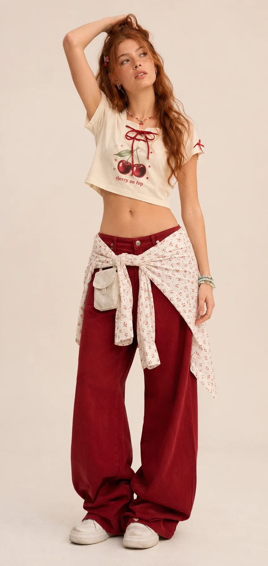 Studio lookbook outfit with a cropped cream graphic tee, cherry-red wide-leg pants, waist-tied printed layer, bracelets, and white chunky sneakers.