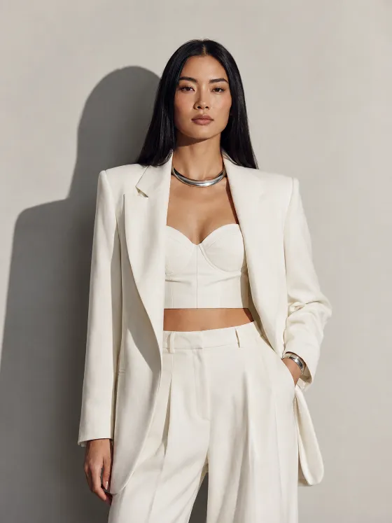 Fashion portrait of an adult woman in ivory tailoring with silver jewelry and a graphic side-cast shadow on a pale gray studio wall.