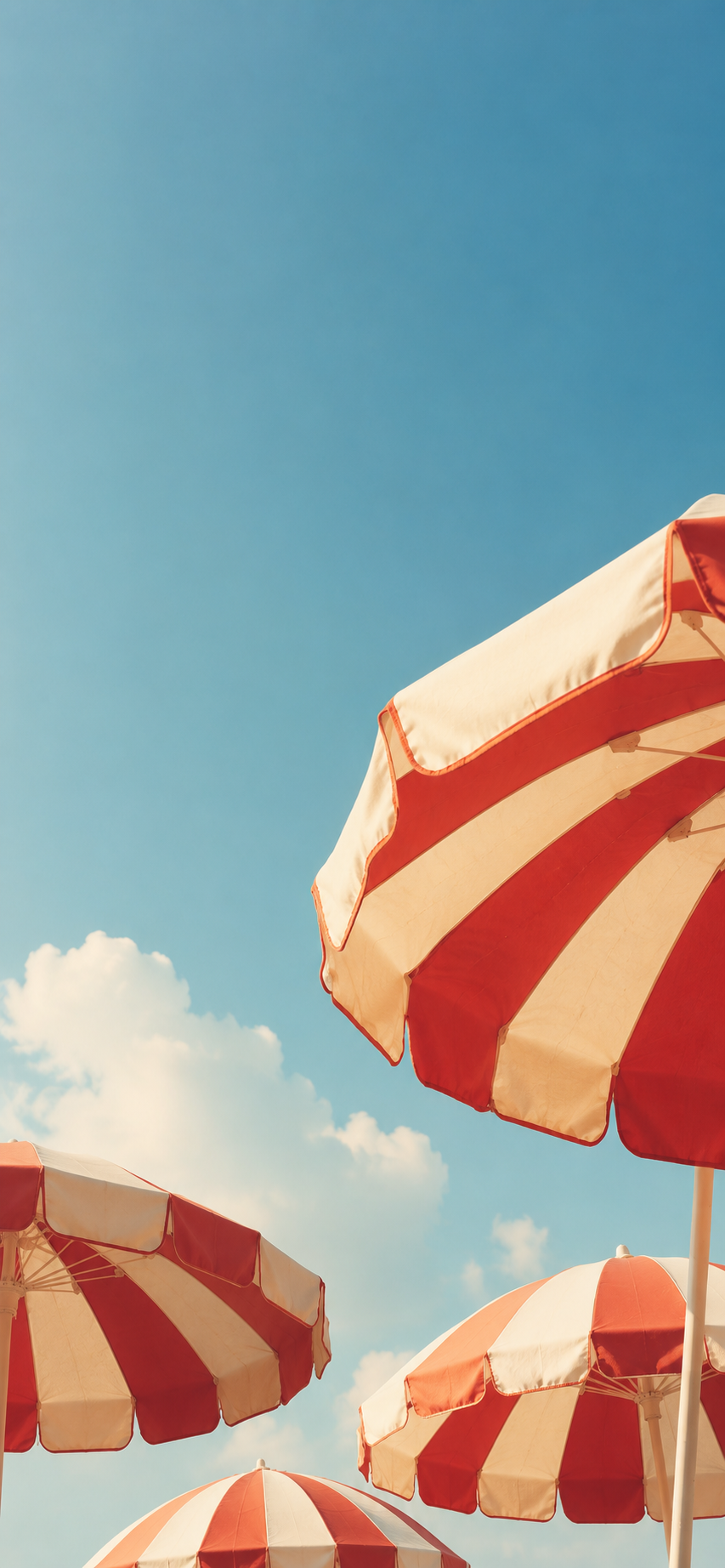 Red and cream striped beach umbrellas framing a bright blue summer sky with soft white clouds.