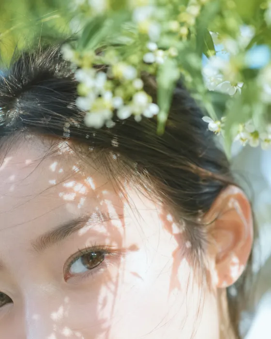 Extreme close-up smartphone portrait of a young woman partly hidden by white flowers and green leaves, with one eye in focus and dappled sunlight on dewy skin.