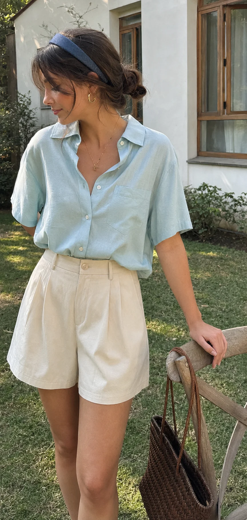 Young woman in a sunlit garden wearing an aqua shirt, cream shorts, and blue headband, standing beside a wooden chair with a woven basket.