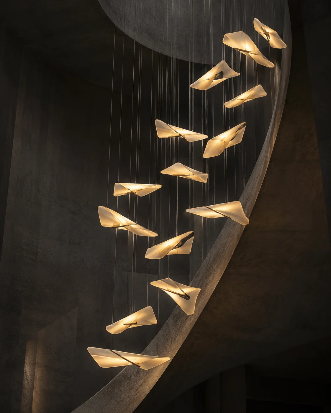 A cluster of glowing wing-like pendant lights hanging beside a large dark curved interior wall.