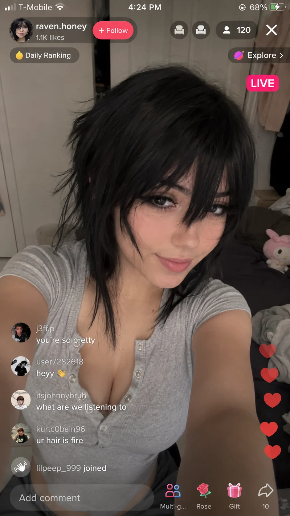 A young woman with short messy black hair and a light gray button-up top takes a selfie in a bedroom with a bed and plush toy visible, TikTok Live int