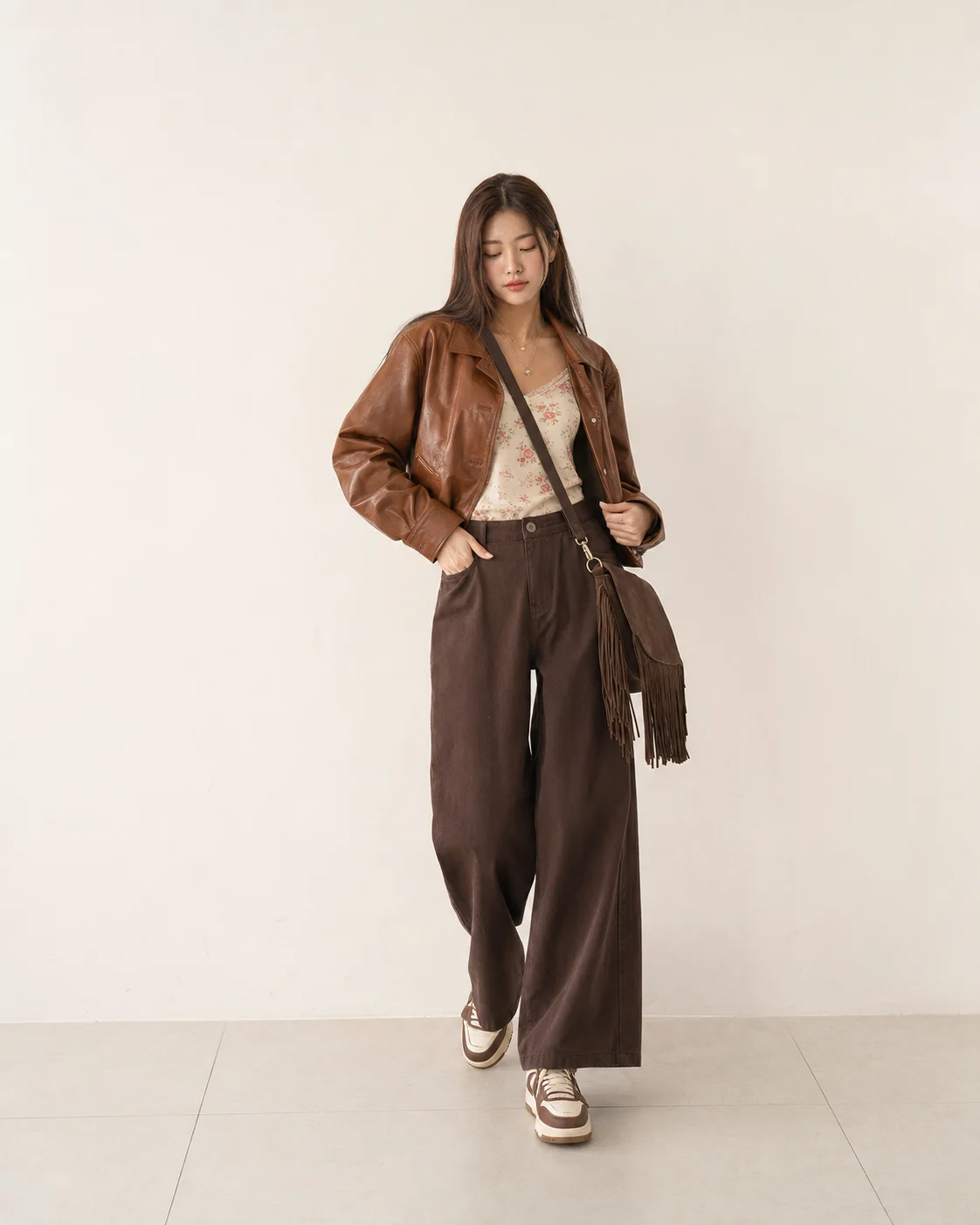 Full-body brown fashion look with a cropped leather jacket, floral ivory camisole, wide-leg trousers and a fringed crossbody bag.