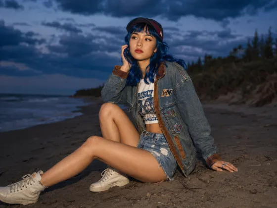 Seated beach fashion portrait with layered denim, cobalt-blue hair, ivory sneakers, dark sand and a cloud-filled twilight sky.