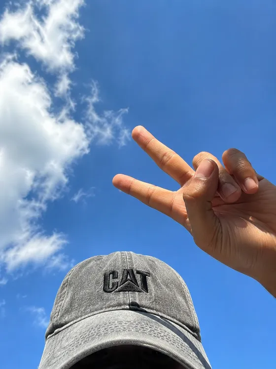 Raised two-finger hand against a bright blue sky with white clouds and a gray CAT baseball cap.