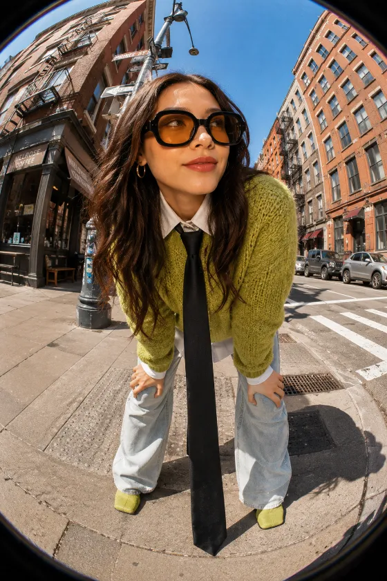 Ultra-wide editorial fashion portrait in a downtown street with green knit styling and oversized sunglasses.