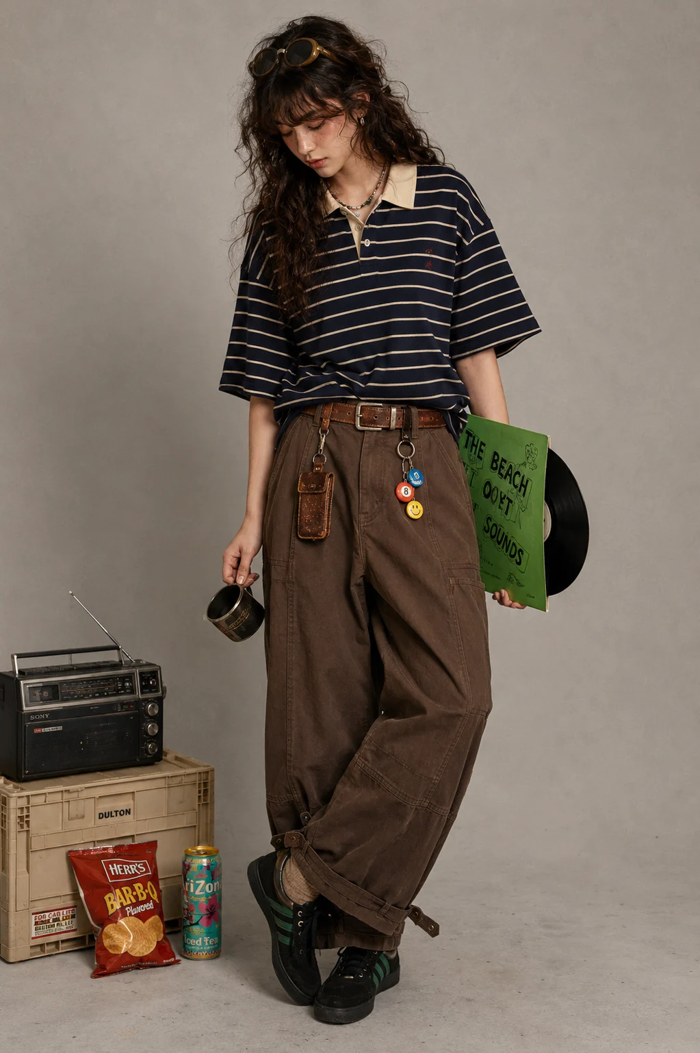 Fashion subject in a navy striped polo and wide brown trousers holding a green record sleeve in a gray studio with vintage props