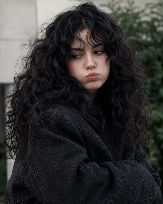 Close outdoor portrait of a woman with voluminous black curls and a puffed-cheek pout, wearing an oversized charcoal jacket in soft overcast light.