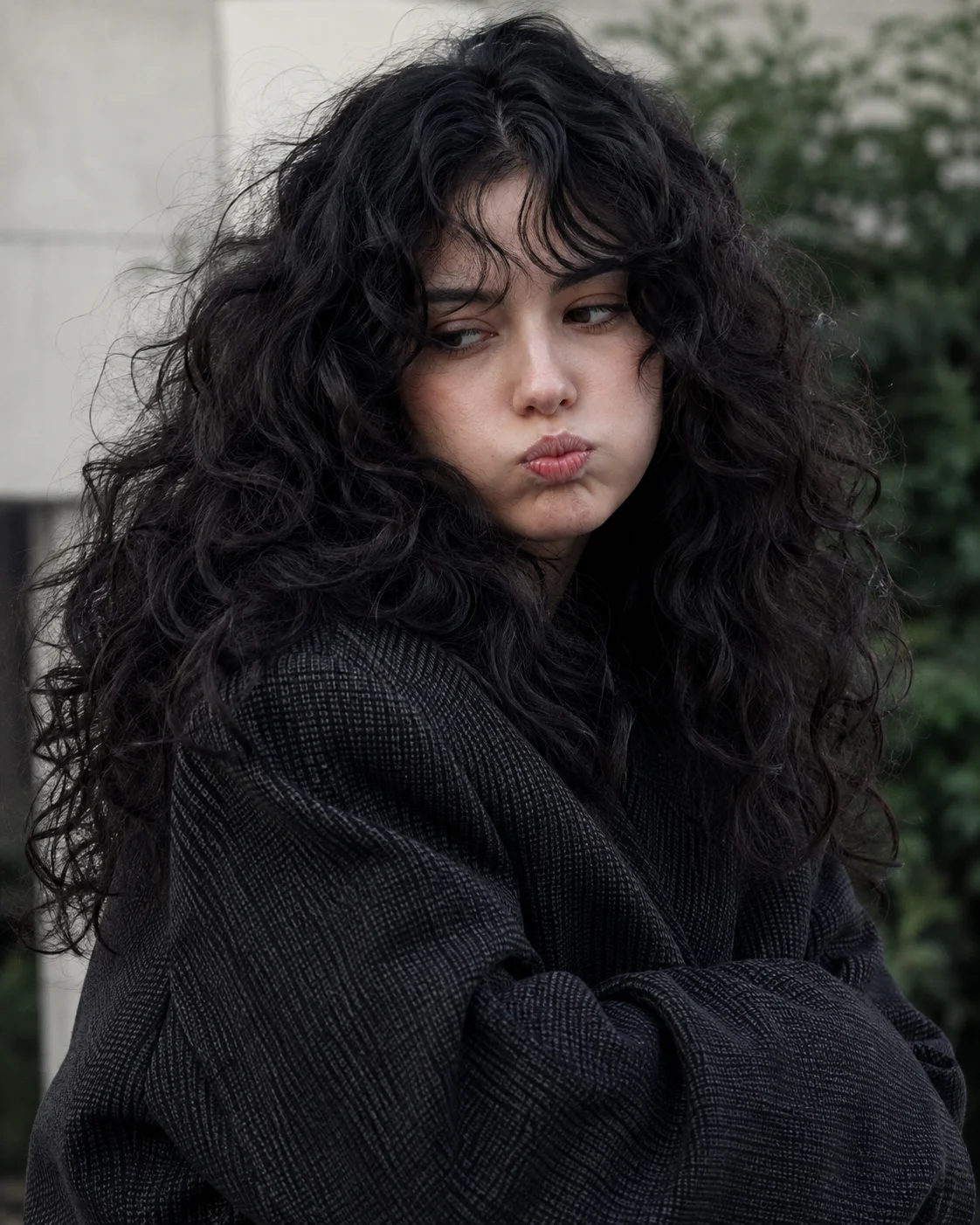 Close outdoor portrait of a woman with voluminous black curls and a puffed-cheek pout, wearing an oversized charcoal jacket in soft overcast light.