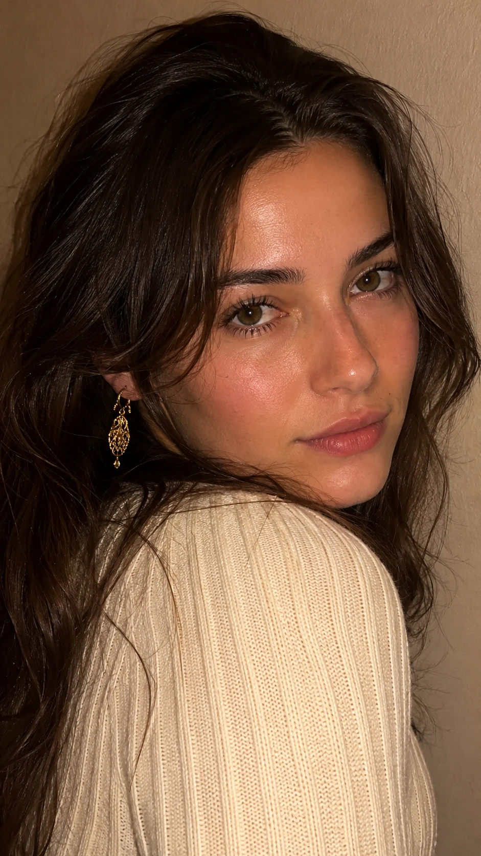 Close-up vertical smartphone portrait of a young brunette woman in a cream ribbed sweater with a gold dangling earring against a warm beige wall.