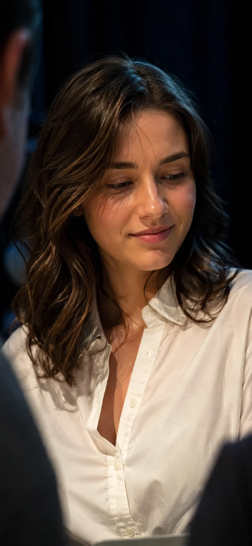 Vertical smartphone portrait of a young brunette woman in a white collared shirt, smiling softly while looking off-camera in warm indoor light.