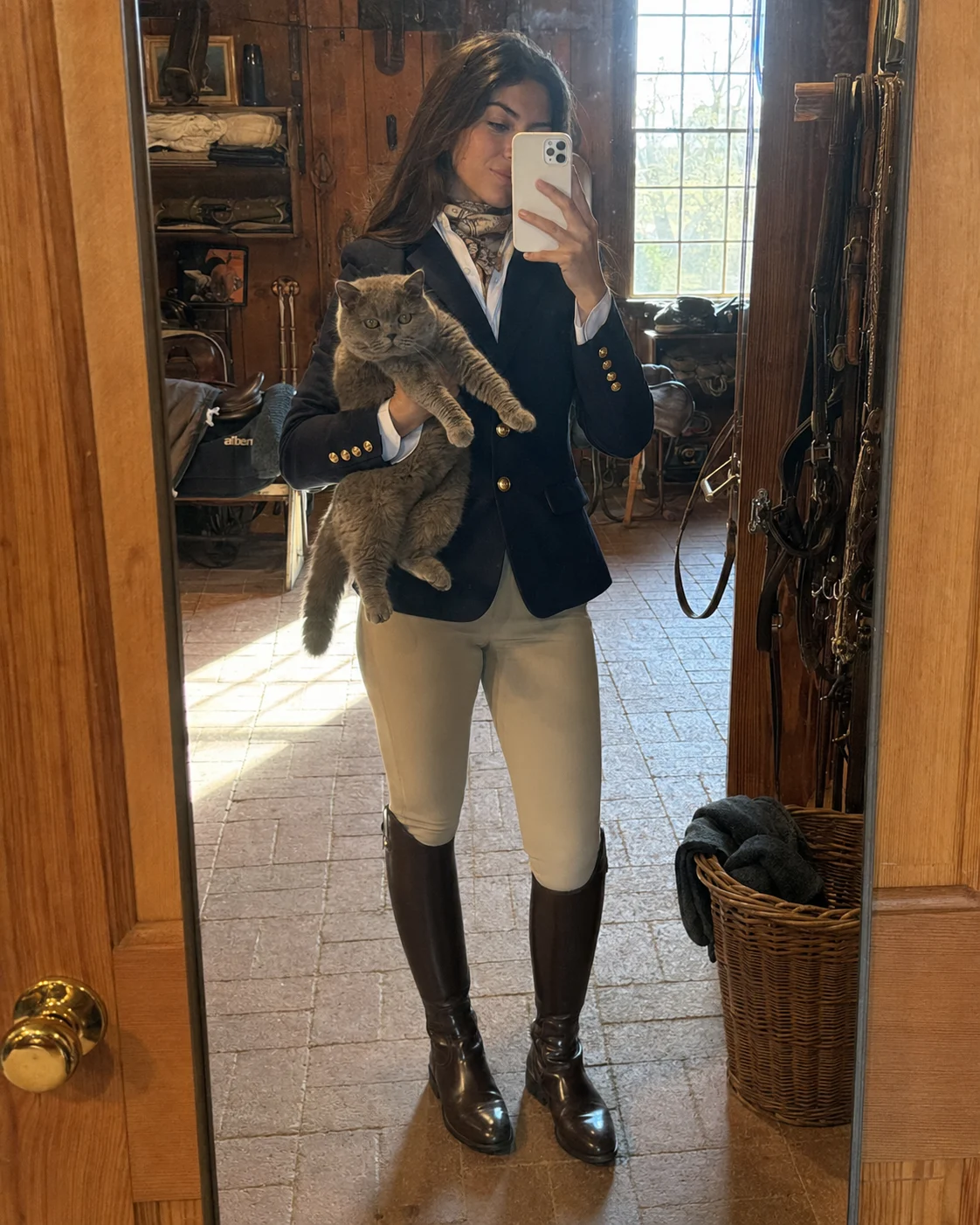 Equestrian mirror selfie in a wooden tack room with navy blazer, beige breeches, tall brown riding boots, scarf, and British Shorthair cat.