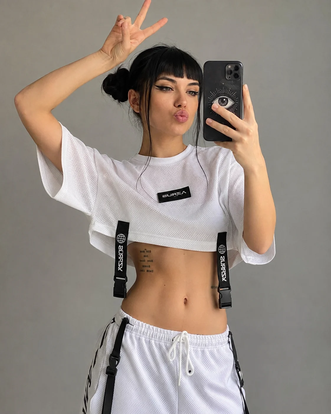 One woman takes a mirror selfie in a white mesh crop top and matching track pants with black straps, buckles, and graphic patches.