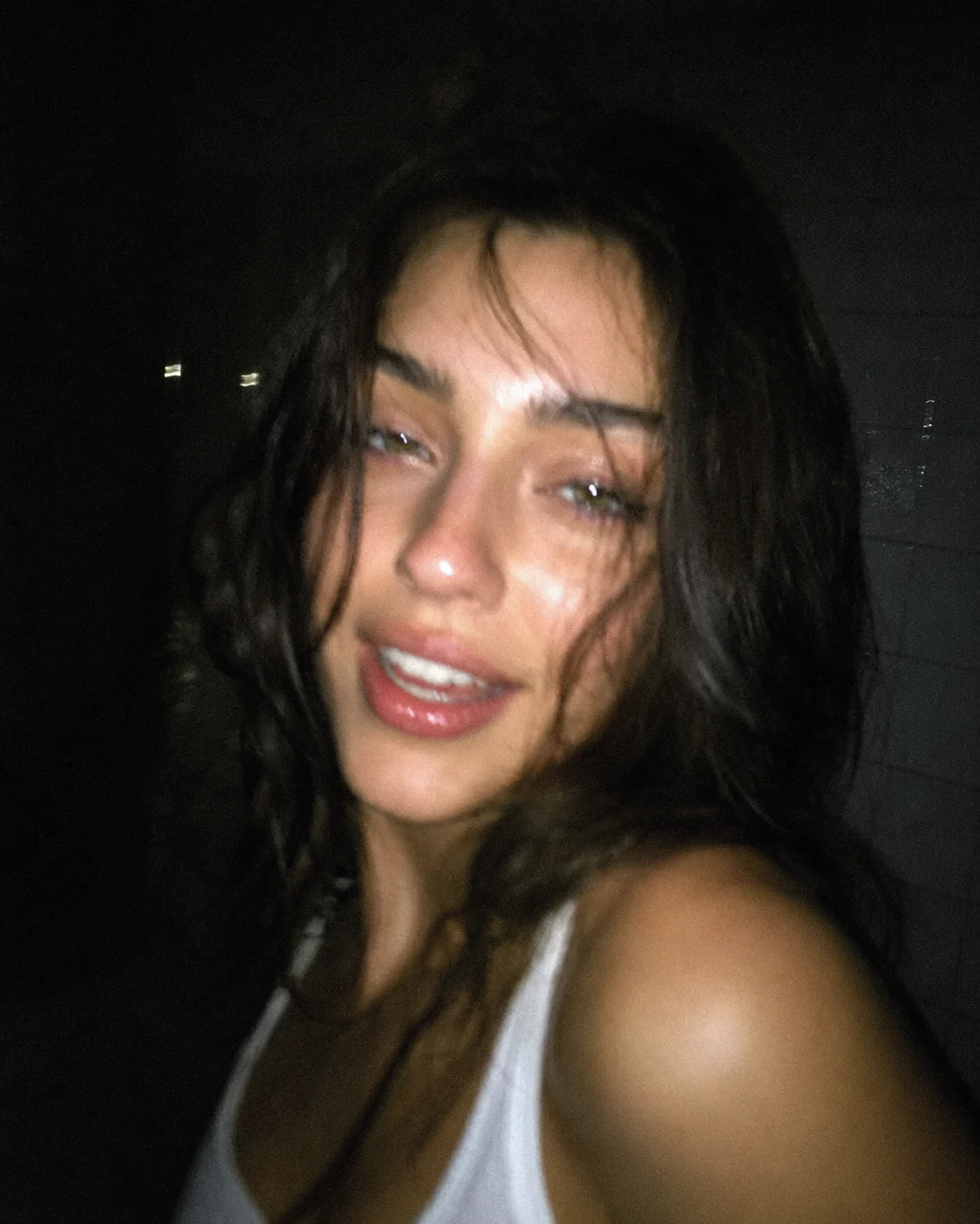 Young woman in a white tank top poses in a dark close-up flash portrait.