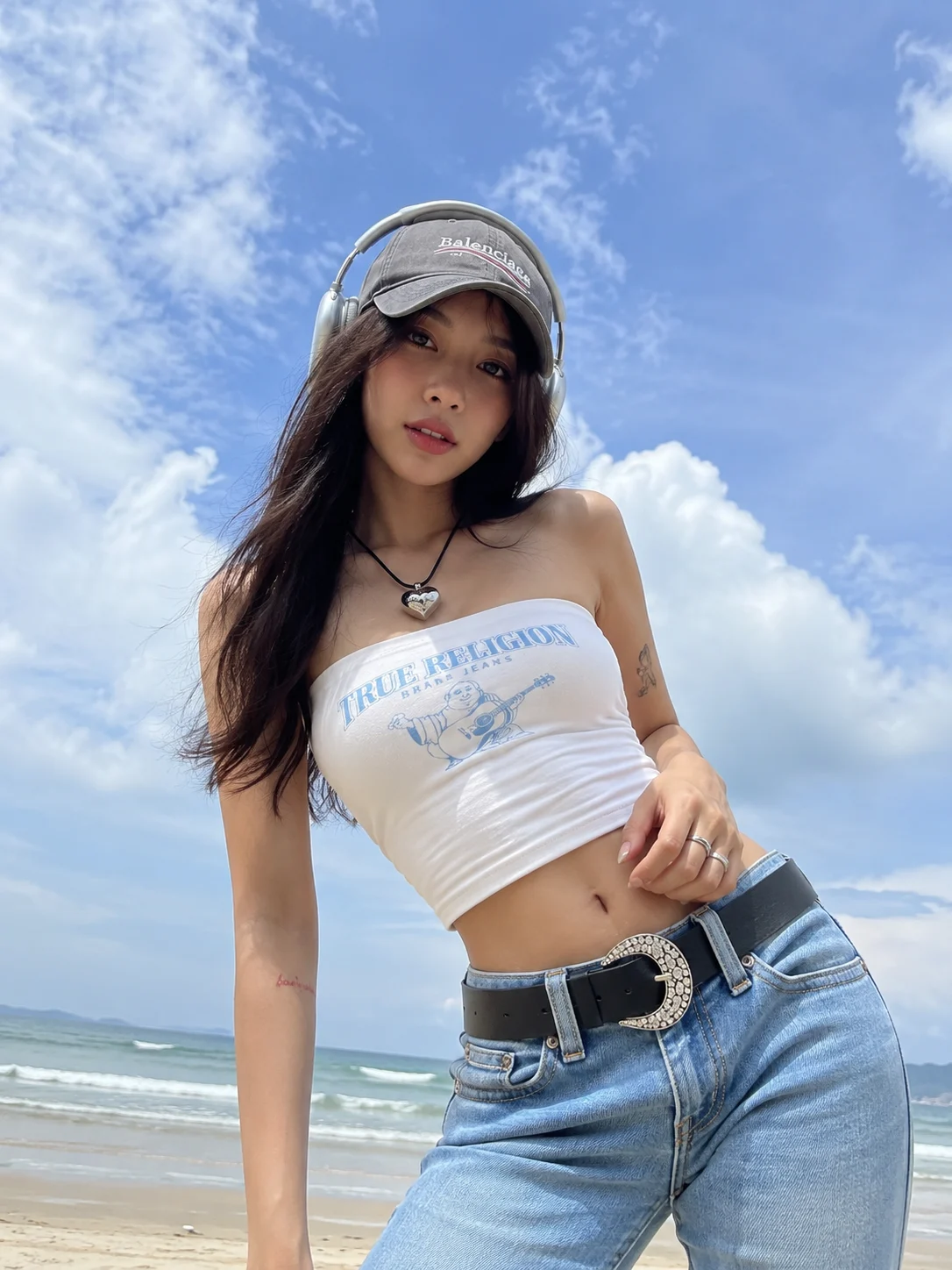 Woman in a casual beach portrait stands near dark seaside rocks with turquoise ocean and clear blue sky behind her, shown as a realistic smartphone-style photo.