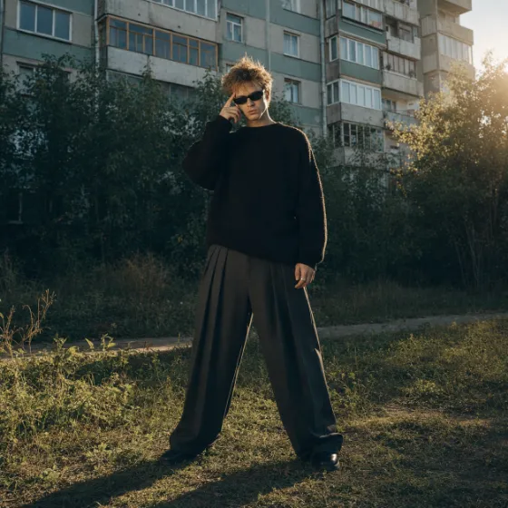 Full-body portrait of a young man in oversized dark menswear and black sunglasses standing in tall grass before shadowed trees and a concrete apartmen