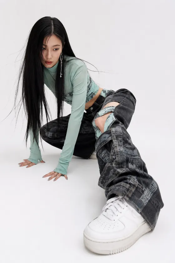 Fashion subject crouched on a white studio floor in a mint long-sleeve top, printed charcoal denim, and white sneakers