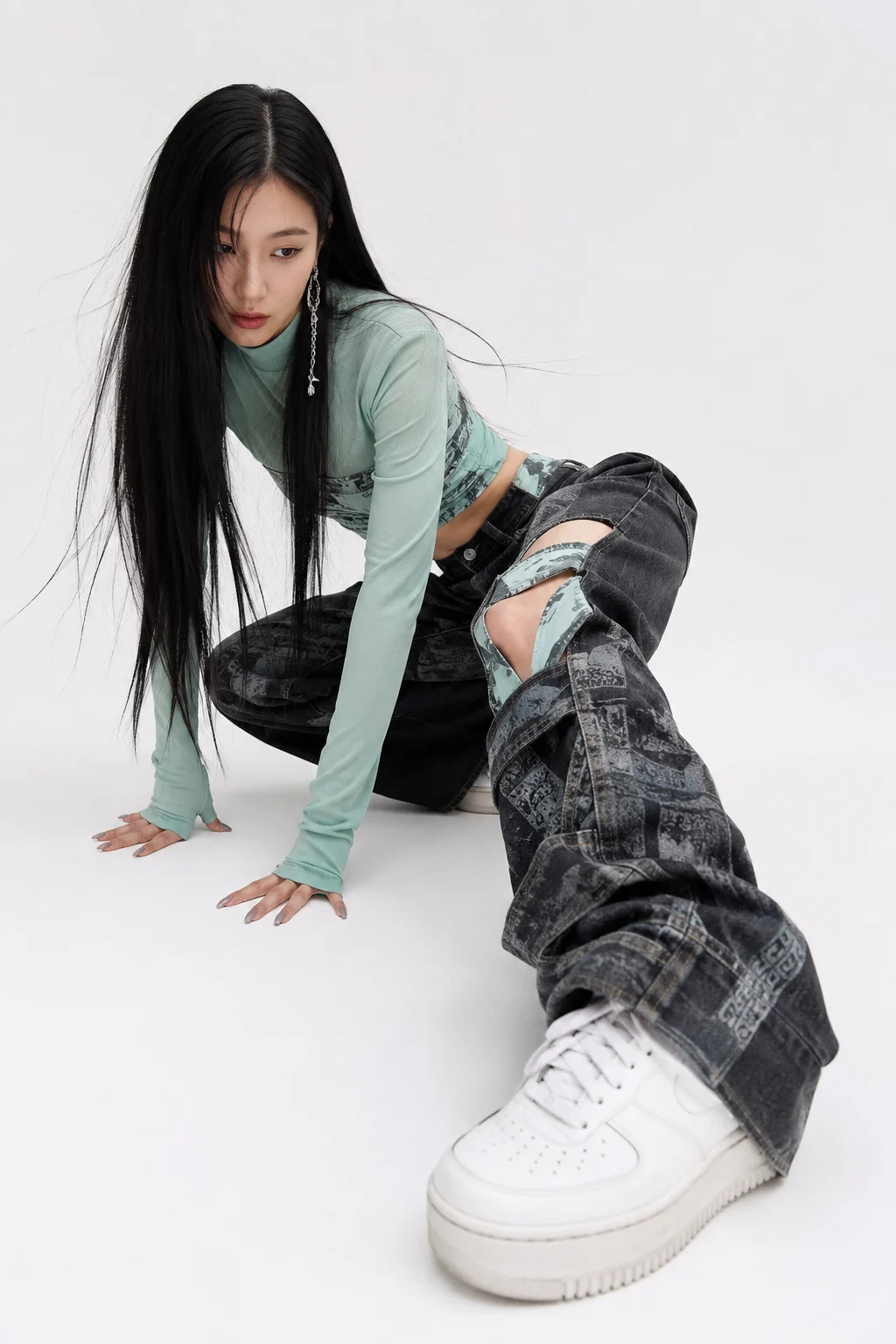 Fashion subject crouched on a white studio floor in a mint long-sleeve top, printed charcoal denim, and white sneakers