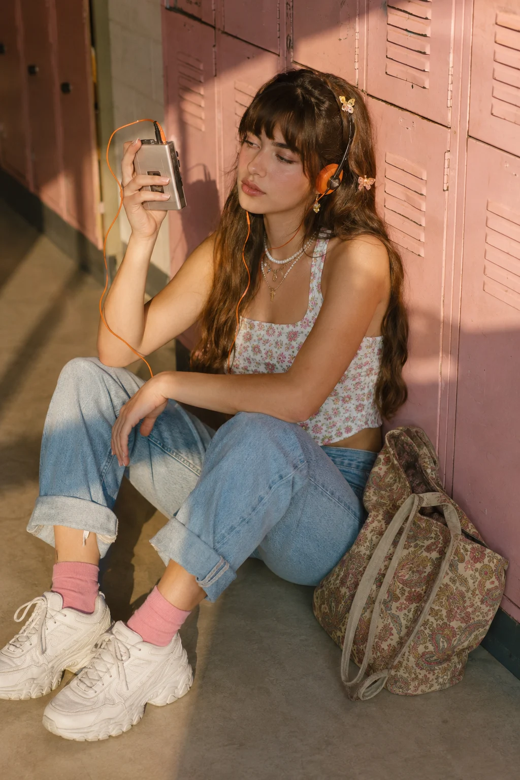 Woman seated by pink lockers in a floral crop top, rolled cuffed jeans, bright pink socks, chunky white sneakers, butterfly clips, and orange headphones.