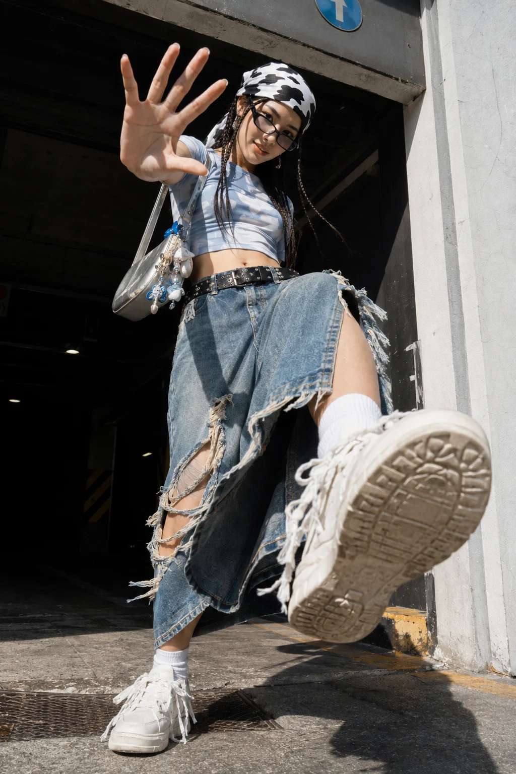 Low-angle urban fashion portrait of a woman in layered denim, a cow-print scarf, metallic bag, and chunky white sneakers.