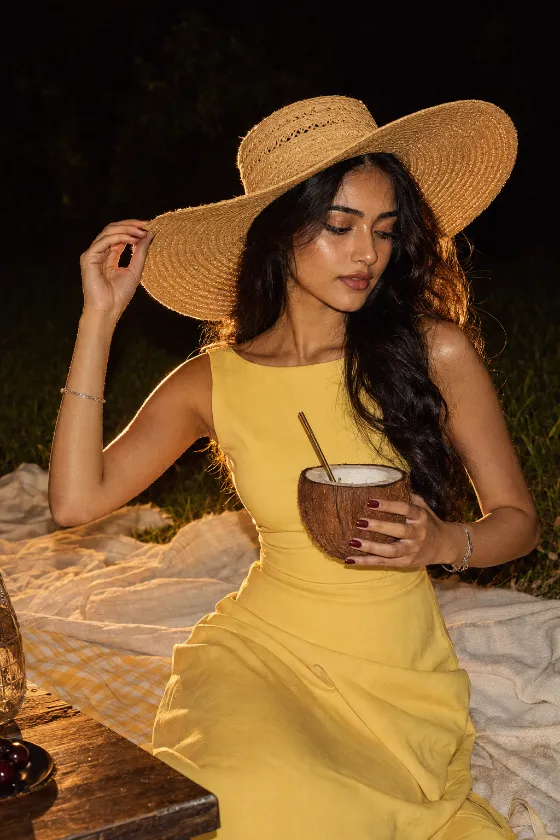 Vertical fashion portrait of a model in a yellow dress and straw hat at a nighttime picnic.