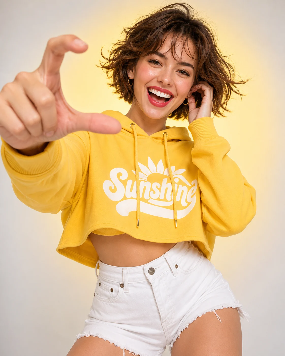 Bright studio fashion portrait with yellow hoodie, white denim shorts, joyful smile, and playful foreground hand gesture.