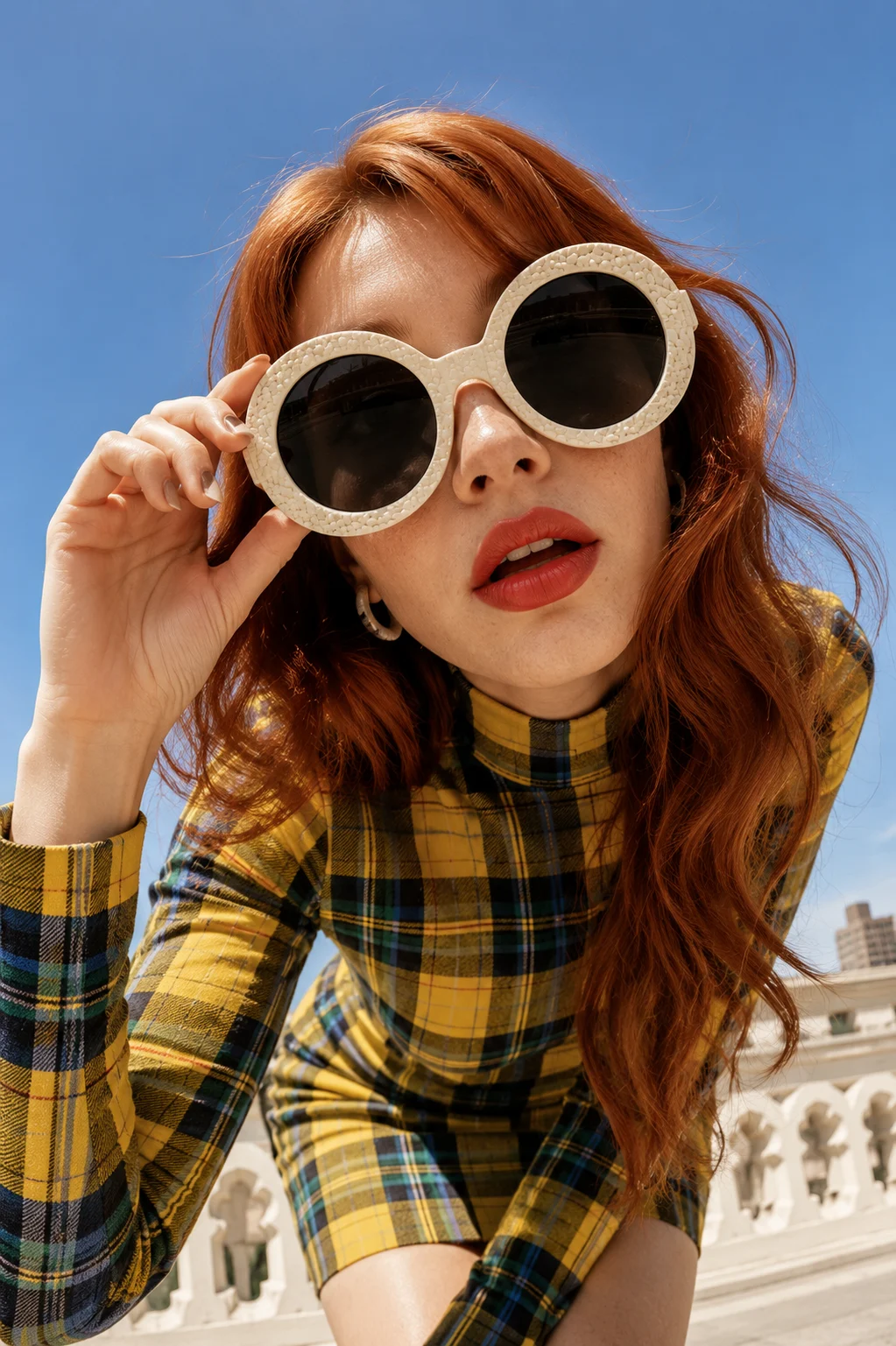 Close editorial of a fashion subject wearing oversized round sunglasses and a yellow plaid outfit outdoors beneath a blue sky.